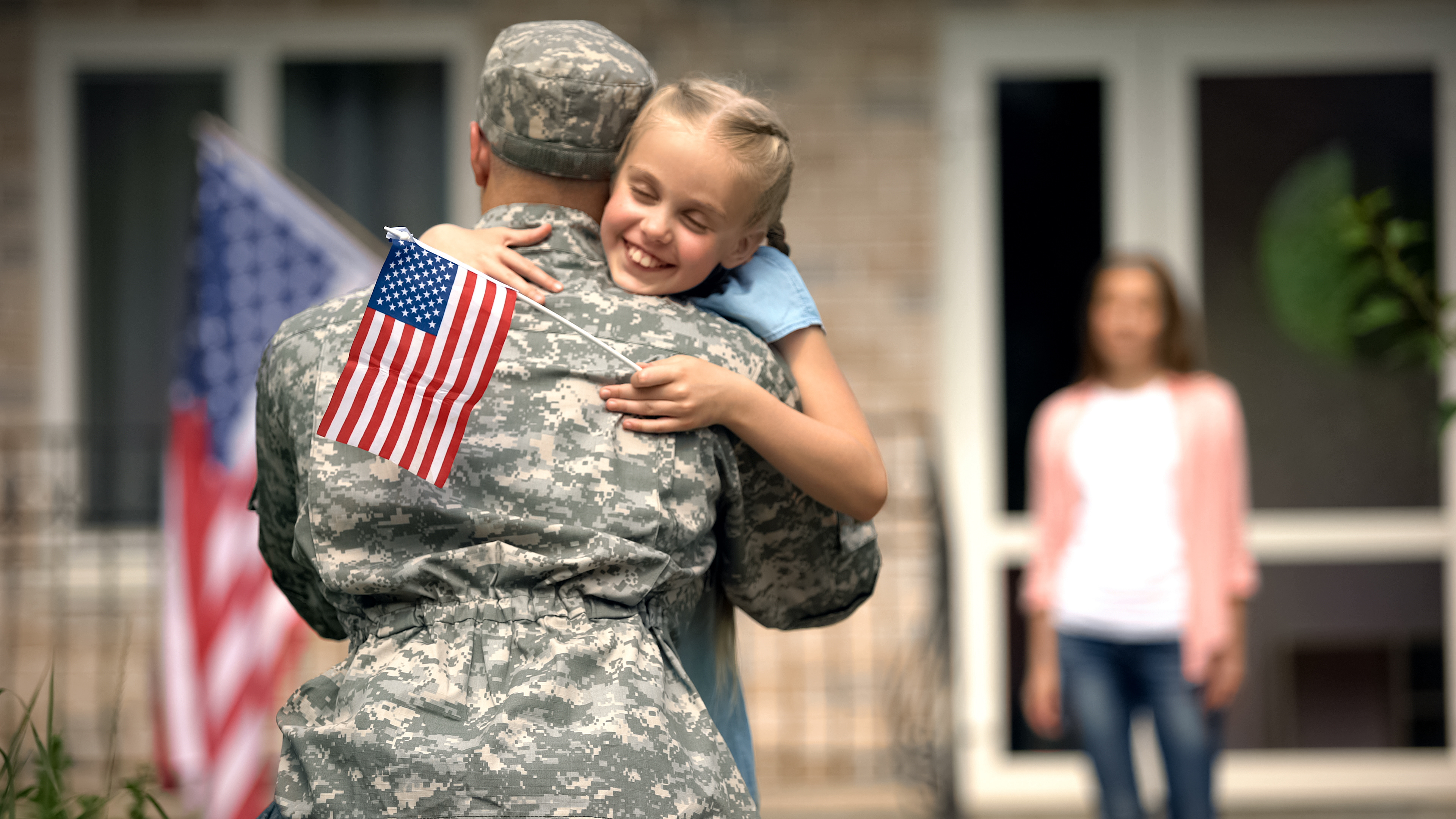 US military service member in camouflage uniform holding young child who is smiling and holding American flag